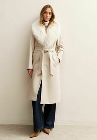 Abrigo beige claro con cuello de piel sintética, detalle de cinturón, mangas largas y bolsillos laterales, combinado con vaqueros oscuros de pierna ancha y tacones puntiagudos en color tostado.