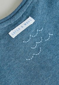 Tissu bleu avec une étiquette blanche cousue portant l'inscription "CATCH A WAVE" et des motifs de vagues brodés en bleu clair à côté de l'étiquette.
