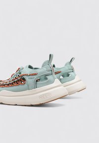 Zapatillas de deporte azul claro con acentos naranjas, parte superior de tela y suela blanca gruesa. Las características incluyen un talón con bucle y un diseño de cordones intrincado.