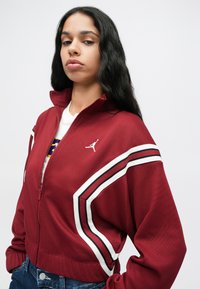 Czerwona kurtka zapinana na zamek z białymi i czarnymi paskami, ściągacze przy mankietach oraz małym białym logo. Nosi się ją na białą koszulę i dżinsy.