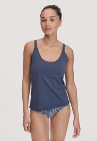 Junge Frau in einem marineblauen ärmellosen Oberteil mit Spitzenrand und passender blauer und grauer gestreifter Unterwäsche vor einem schlichten Hintergrund.