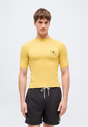 Młody mężczyzna w dopasowanym żółtym rash guardzie z logo i czarnych krótkich kąpielówkach z białym troczkiem, stojący na białym tle.