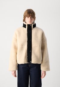Beige nep-bontjasje met een zwarte nylon voorkant, drukknoopsluitingen, brede mouwen en een hoge kraag. Draagt over donkere spijkerbroeken.