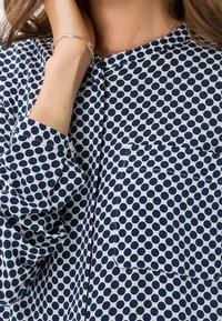 La camicia a pois blu e bianca presenta maniche lunghe, un collo rotondo e una tasca sul petto. Tessuto morbido con una consistenza liscia.