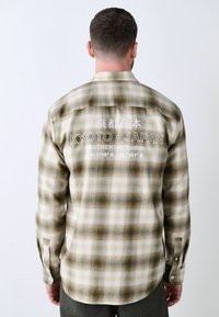 Camicia in flanella a quadri beige e verde oliva, con una grafica sul retro che presenta "Kyoto, Giappone" e le coordinate. Tessuto leggero, maniche con bottoni.