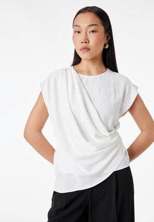 Nainen, jolla on pitkät mustat hiukset, hopeiset renkaankorvakorut ja valkoinen liehuva hihaton paita, seisoo käsivarret lanteilla valkoista taustaa vasten.