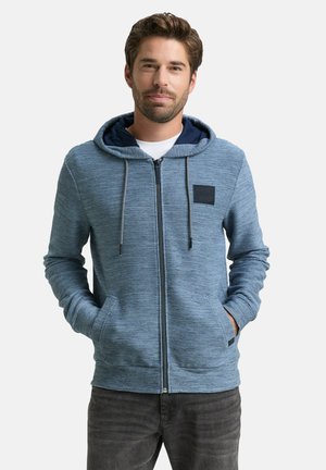 Mann trägt einen blauen Zip-Hoodie mit gestreifter Textur und Kordeln, Hände in den Taschen, steht vor einem schlichten weißen Hintergrund.