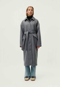 Grå lang frakke med beltestrop om livet, knaplukning foran og stor krave. Båret over en sort turtleneck og lyseblå flare jeans med sneakers.