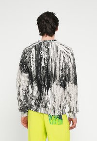 Hosszú ujjú pulóver fekete-fehér absztrakt mintával, bordázott mandzsettákkal és szegéllyel. Élénksárga nadrággal párosítva.