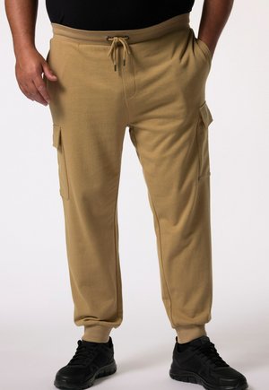 Mand iført beige cargo jogger-bukser med snøre i taljen og sorte sneakers, stående med hænderne nær lommerne på hvid baggrund.