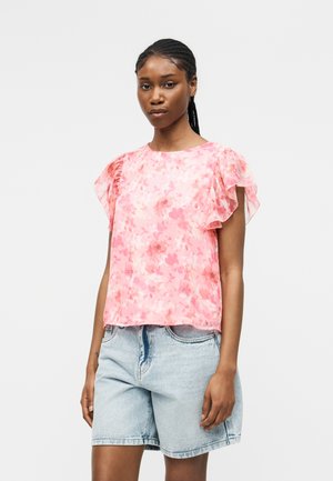 Kobieta z plecionymi włosami, ubrana w różową kwiecistą bluzkę z falbankami na rękawach oraz jasne dżinsowe szorty, stojąca na jednolitym tle.