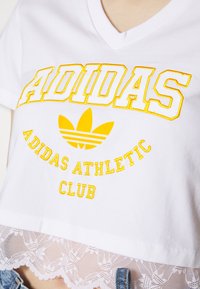 Balta medvilninė marškinėlių su geltonu siuvinėtu "ADIDAS" tekstu ir logotipu. Savybės: nėriniai apatine dalyje ir V formos kaklo dizainas, trumpomis rankovėmis.
