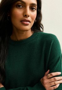 Zielony sweter z dzianiny, z okrągłym dekoltem. Tkanina wygląda na miękką, z fakturowanym wyglądem. Złote kolczyki w kształcie kół oraz pierścionki dopełniają dodatki.