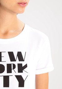 Camiseta blanca de algodón con un texto negro en negrita que dice "NUEVA YORK," cuello redondo y mangas cortas enrolladas.