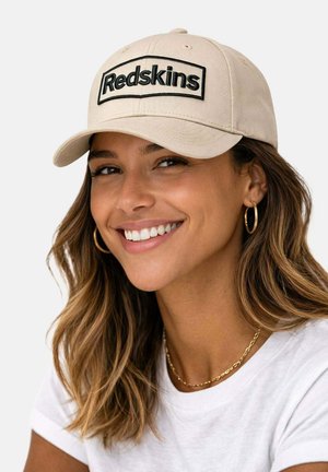 Femme souriante aux longs cheveux ondulés portant des boucles d'oreilles créoles dorées, un collier en or, une casquette beige avec le texte « Redskins » et un t-shirt blanc.
