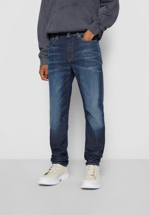 Jeans de mezclilla azul con corte recto, ligeras desgastadas en los muslos, cinco bolsillos y un diseño clásico de trabillas, combinados con zapatillas beige.
