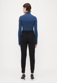 Suéter de punto azul con cuello alto combinado con pantalones negros ajustados. Presenta una cintura elástica y bolsillos traseros. La modelo lleva zapatos de tacón negros.