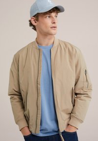Beige bomber dzseki, cipzárral, bordázott mandzsettákkal és oldalsó zsebekkel, egy világoskék ing fölé rétegezve. A világoskék sapka teszi teljessé a megjelenést.