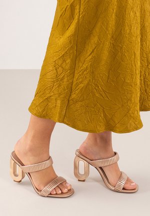 Sandalias de tacón beige con tiras texturizadas, punta abierta y un diseño de tacón geométrico único. Combinadas con una falda arrugada mostaza.