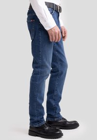 Férfi kék farmernadrágban, fekete bőrcipőben és fehér hosszú ujjú ingben, világosszürke háttér előtt állva.