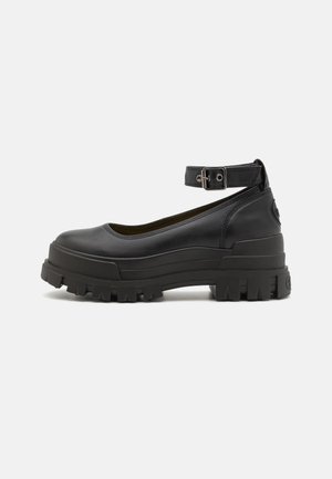 Chaussure plateforme noire épaisse avec semelle à crampons épaisse et bride de cheville ajustable avec boucle en métal, vue de profil sur un fond uni.