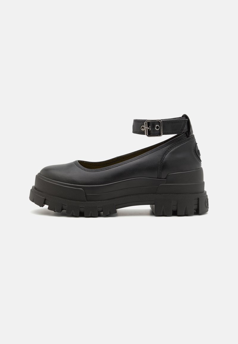 Chaussure plateforme noire épaisse avec semelle à crampons épaisse et bride de cheville ajustable avec boucle en métal, vue de profil sur un fond uni.