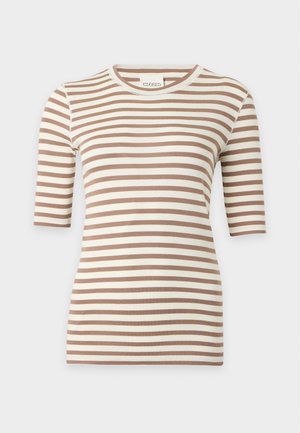 Femme aux longs cheveux foncés portant une chemise beige et blanche rayée et un jean large bleu foncé en denim, debout sur un fond blanc.