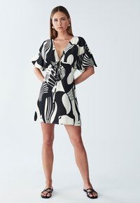 Kurzärmeliges Minikleid in Schwarz und Weiß mit abstrakten Mustern, ausgestattet mit einem V-Ausschnitt und einer Bindung in der Taille. Flache Sandalen.