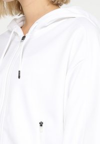 Sweat à capuche blanc à fermeture éclair avec capuche, poches avant zippées et tissu texturé. Comprend un cordon ajustable et des détails en métal contrastants.