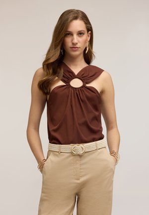 Vrouw met lang golvend haar met een mouwloos bruin topje met een ronde uitsnijding en beige broek met een geweven riem en gouden sieraden.