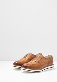 Chaussures en cuir marron avec des perforations et un design de bout ailé ; orteils arrondis, semelles intermédiaires blanches et rayures noires contrastantes.