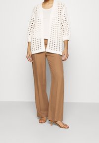Pletený cardigan v odstínu off-white, s otevřeným vzorem a rukávy po tři čtvrtě, doplněný hnědými širokými kalhotami a nahými sandály na pásky.