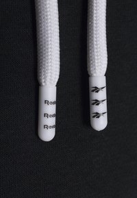 Białe warkoczykowe sznurowadła z plastikowymi końcówkami; jedna końcówka ma czarny napis "Reebok", podczas gdy druga ma wzór logo na czarnym tle.