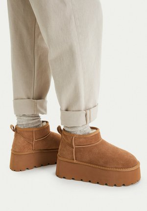 Person wearing light beige cuffed pants, beige textured socks, and brown suede slip-on platform boots with thick soles.