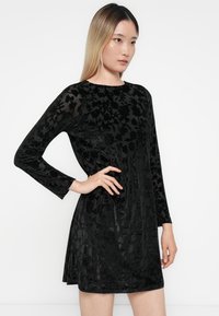 Abito in velluto nero con maniche lunghe, scollo rotondo e texture a motivo floreale, che si allarga all'orlo sopra la metà della coscia.