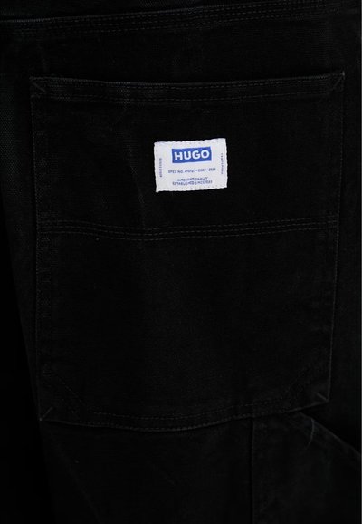 Gros plan sur une poche de vêtement noir, avec une étiquette blanche de la marque HUGO cousue près du haut.