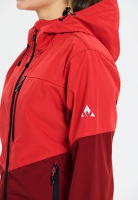 Red jacket with a hood, featuring a two-tone design, black zipper, and logo accent on the sleeve. Smooth texture and athletic cut.