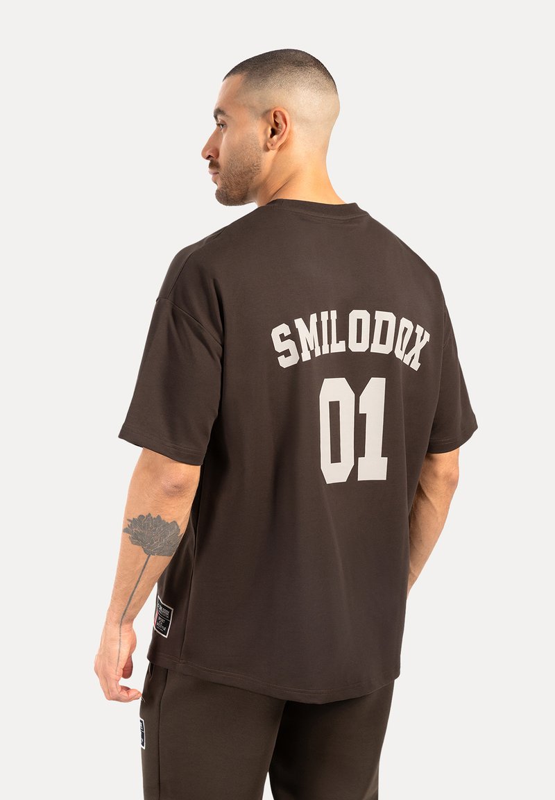 Brun oversized t-shirt lavet af bomuld, med hvidt trykt tekst "SMILODOX 01" på ryggen. Korte ærmer og crew neck-design.