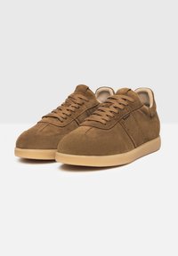 Sneakers in suede marrone con design a basso profilo, lacci piatti e suola in gomma di un beige chiaro. Presentano cuciture laterali e una texture liscia.