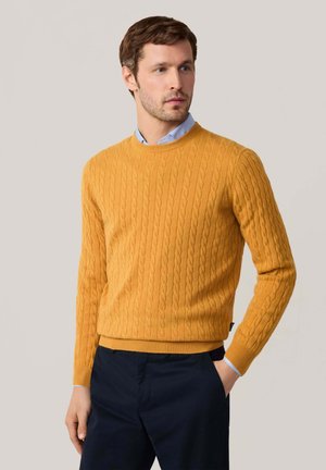 Suéter de punto cable en color mostaza con cuello redondo y mangas largas, combinado con pantalones oscuros. Tejido texturizado y diseño ajustado.