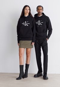 Dva modela nose crne dukserice s bijelim logotipima, uparene sa zelenom suknjom i krojenim crnim hlačama. Oboje nose crnu obuću. Minimalna pozadina.