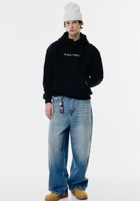 Felpa nera con testo bianco, jeans baggy azzurri chiari, stivali da lavoro color sabbia e berretto di maglia bianco. Dotata di grandi tasche e cappuccio con cordino.
