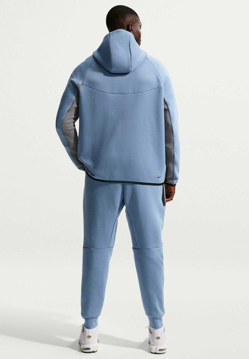 Felpa con cappuccio blu dal taglio comodo e cappuccio regolabile, con dettagli grigi sulle maniche. Abbinata a pantaloni della tuta coordinati. Tessuto strutturato.