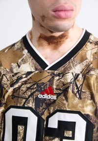 Kamufleret trøje i nuancer af brun og beige, med en sort krave, hvidt trim og sorte tal "03" samt rød Adidas-logo.