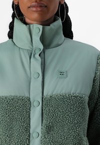 Mäťovo zelená bunda so mäkkou, textúrovanou fleece dolnou časťou, hladkým horným materiálom a gombíkovým zapínaním. Malé logo na hrudi.