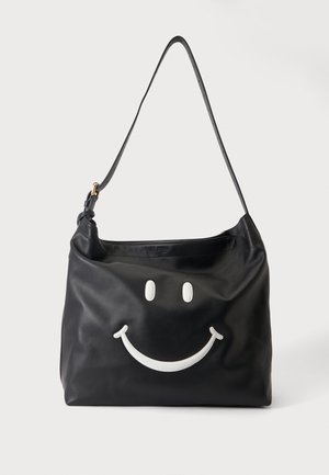 Sac à épaule en cuir noir avec longue bandoulière et motif visage souriant blanc sur le devant sur fond clair.