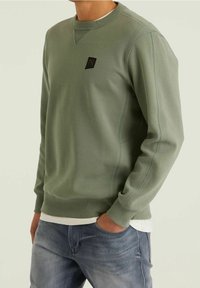 Sweat-shirt vert avec un col rond, des poignets côtelés et des coutures latérales, arborant une petite étiquette de logo noir sur la poitrine. Porté par-dessus une chemise blanche.