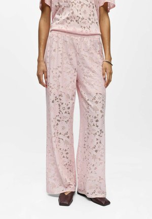 Femme portant un pantalon large en dentelle rose et un haut en dentelle assorti, accompagné de chaussures sombres à bout carré, debout sur un fond blanc.