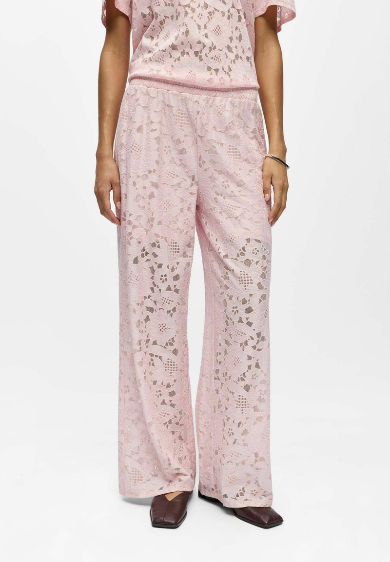 Femme portant un pantalon large en dentelle rose et un haut en dentelle assorti, accompagné de chaussures sombres à bout carré, debout sur un fond blanc.