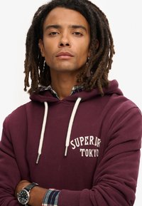 Felpa bordeaux con cordini bianchi e testo "Superdry Tokyo". Sotto, una camicia a quadri con motivi blu, verde e rosso. Orologio nero al polso.